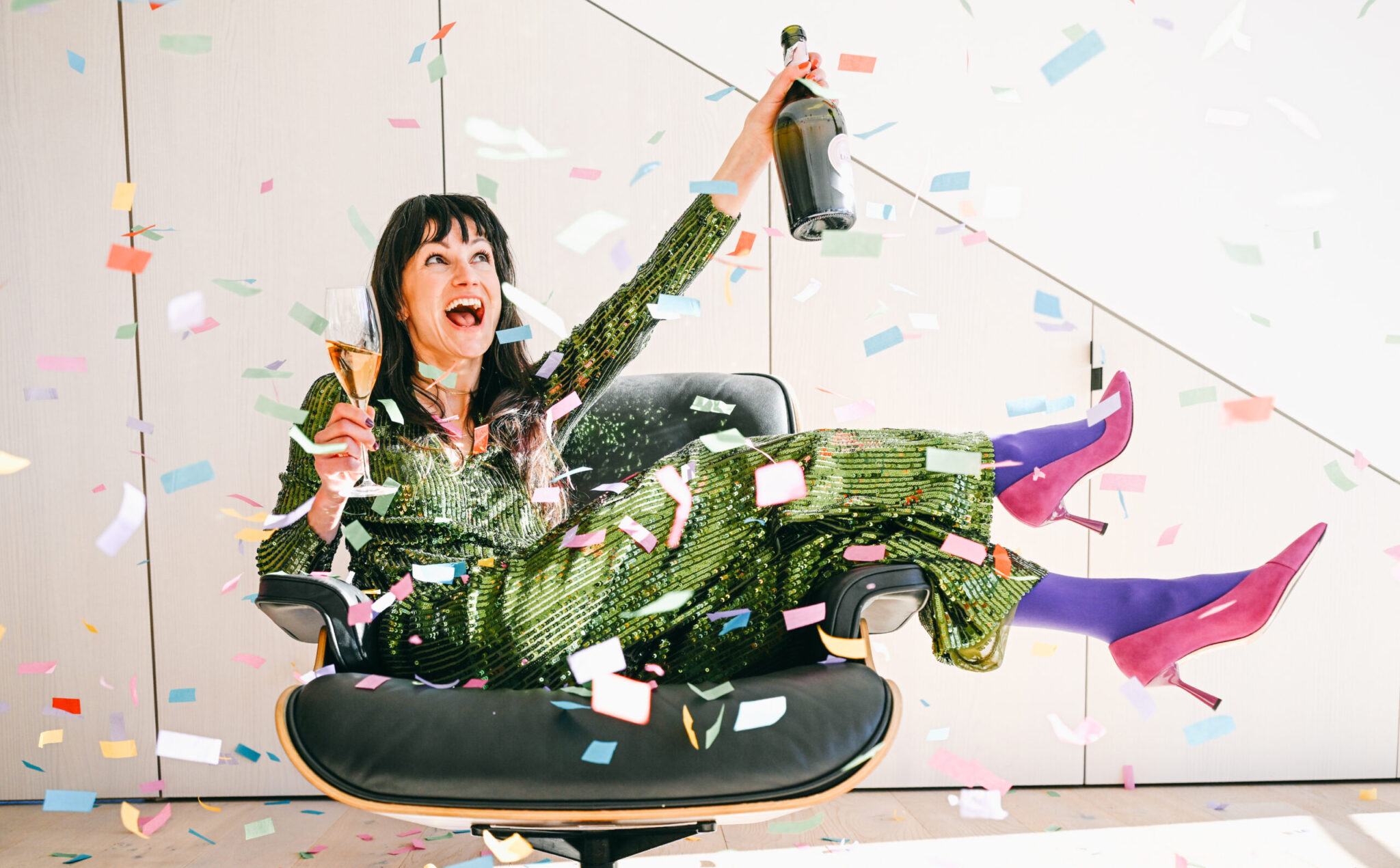 Frau mit einem Sektglas in der Hand sitzt auf einem Sessel in einem Raum mit Konfetti
